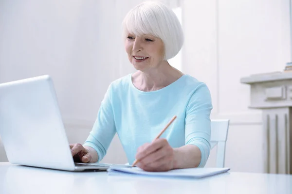 Улыбающаяся пожилая женщина, работающая за ноутбуком — стоковое фото