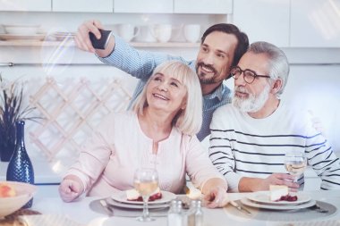 Just smile. Positive delighted adult son takign selfies with his aged parents while sitting together in the kitchen and expressing gladness clipart