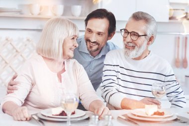 Warm relationships. Cheerful delighted aged couple sitting at the table while his adult son embracing them and resting in the kitchen clipart