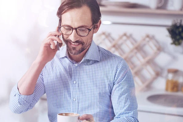 Always in touch. Cheerful delighted adult man sitting in the kitchen and talking on smart phone while drinking coffee