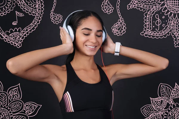 Great sound. Young slim sportswoman looking happy while wearing modern convenient headphones and listening to pleasant music