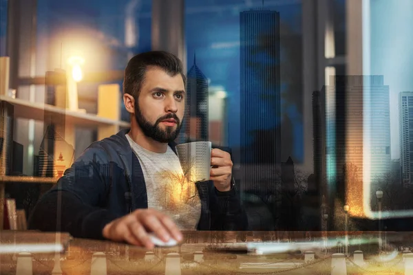 Uppmärksamma mannen håller en kopp medan du arbetar på framsidan av hans dator — Stockfoto