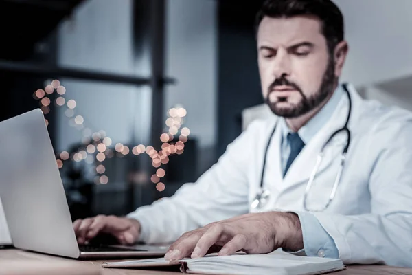 Médico confiante ocupado usando laptop e olhando para seu caderno . — Fotografia de Stock