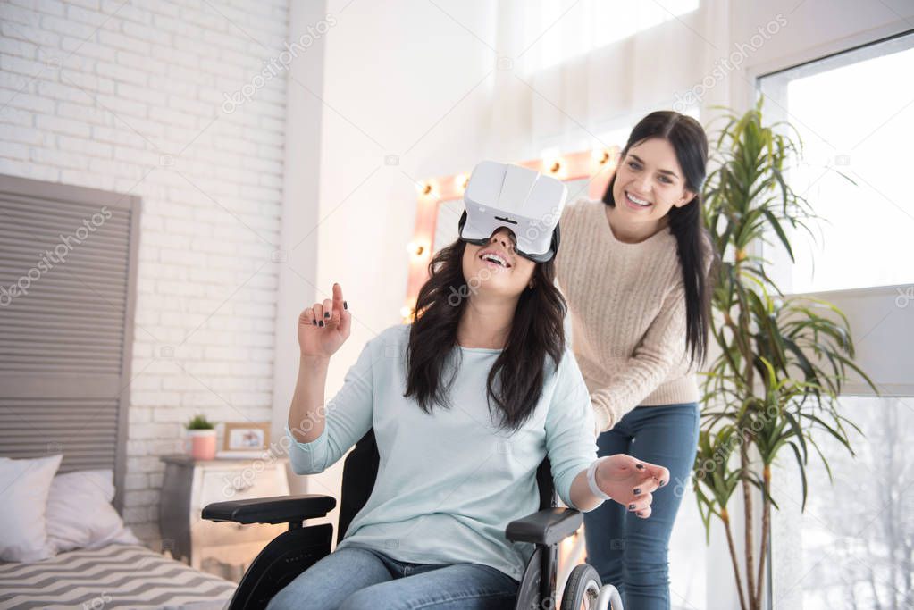 Virtual world. Merry friend staying while immobile woman wearing VR glasses