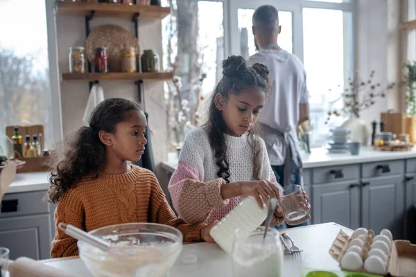 Irmã derramando leite em vidro enquanto cozinha torta com a irmã — Fotografia de Stock