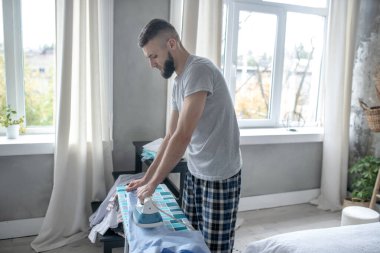 Sakallı adam pencerenin yanında duruyor ve kıyafetleri ütülüyor.