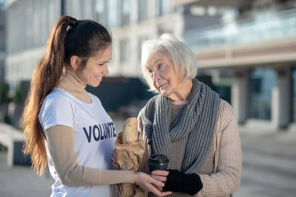 Volunteer supporting poor woman while bringing bread and tea — Stock Photo, Image