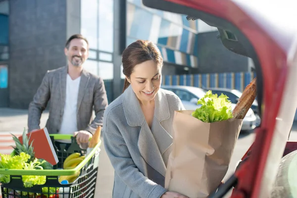 Жена улыбается, когда кладет сумку с едой в машину после покупок — стоковое фото