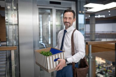 Smiling successful businessman holding box while standing near elevator clipart