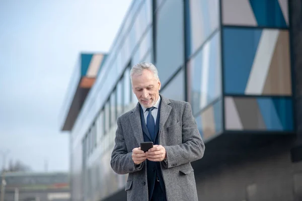 Сіроволоса людина в сірому пальто перевіряє свій телефон — стокове фото