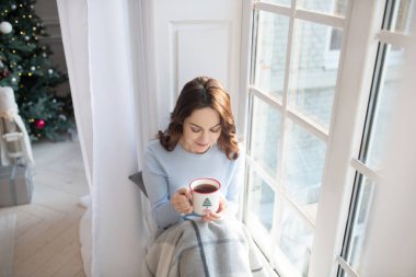 Koyu saçlı, gri gömlekli genç bir kadın pencerenin yanında kahve içiyor.