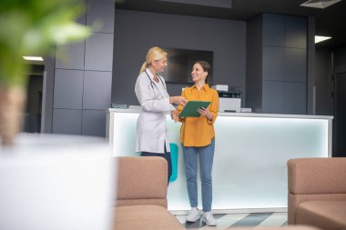 Sarışın doktor beyaz cübbeli hastayla konuşuyor.
