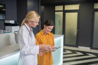 Sarışın doktor hastanın sağlık raporunu doldurmasına yardım ediyor.