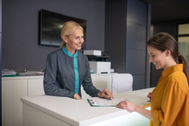 Klinikteki sarışın resepsiyonist bir ziyaretçiyle konuşuyor.