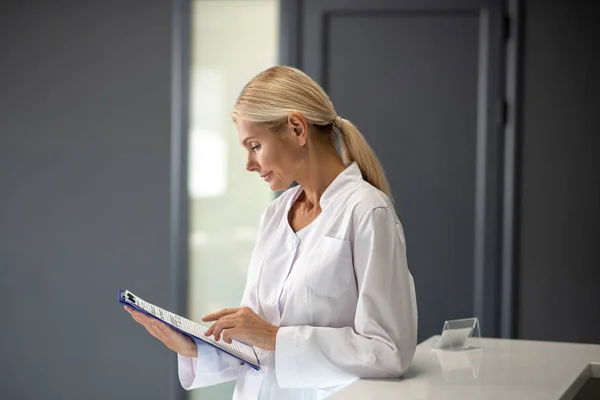 Blond vacker läkare i vit rock tittar igenom uppdragsblad — Stockfoto