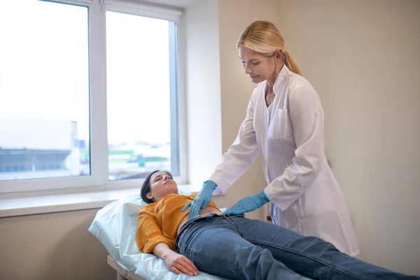 Steril eldiven takan sarışın doktor hastaların midesine dokunuyor. — Stok fotoğraf