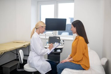 Sarışın doktor, genç siyah saçlı hastasıyla konuşuyor.