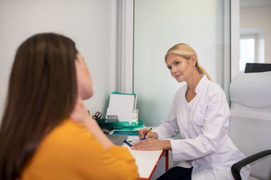 Beyaz cüppeli sarışın doktor muayene sonuçlarını hastaya söylüyor.