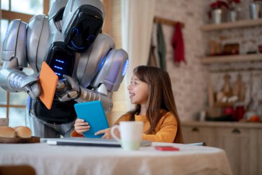 Robot turuncu gömlekli kıza bir şey gösteriyor.