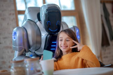 Turuncu tişörtlü gülümseyen kız robotla selfie çekiyor.