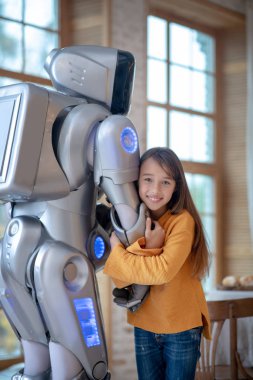 Uzun saçlı güzel kız ev robotuna sarılıyor.