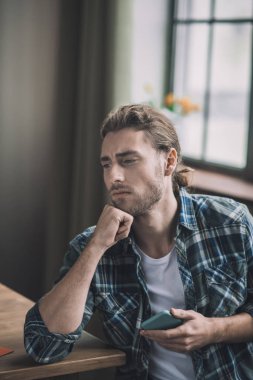 Endişeli uzun saçlı adam akıllı telefonunu elinde tutuyor.