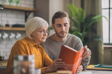 Zarif görünümlü gri saçlı kadın ve oğlu kitap okuyorlar.