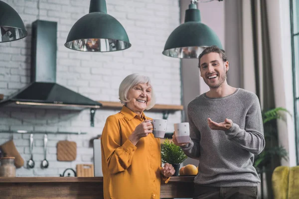Jonge knappe man in grijs shirt en zijn moeder praten in de keuken — Stockfoto