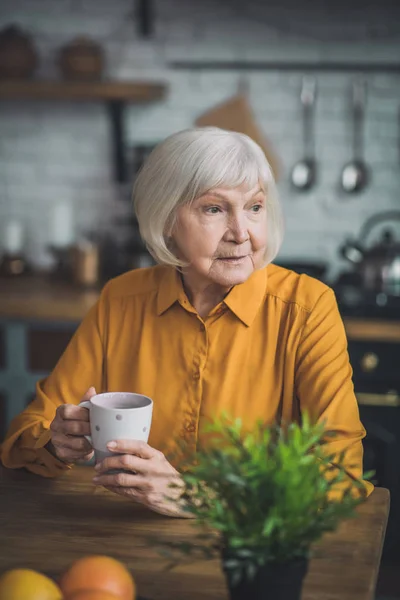 Sarı elbiseli güzel yaşlı bayan mutfağında oturuyor. — Stok fotoğraf