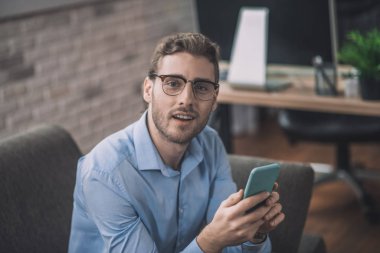 Mavi gömlekli genç sakallı adam akıllı telefonunu tutuyor.