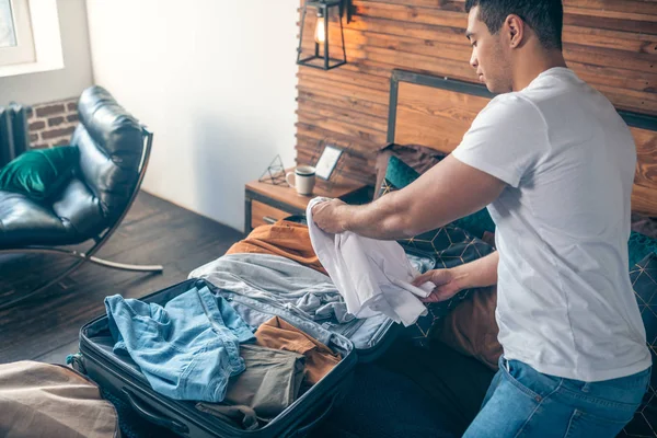 Joven de cabello oscuro con camiseta blanca ocupado empacando ropa —  Fotos de Stock