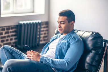 Elinde akıllı telefonuyla sandalyede oturan genç adam..