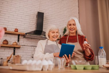 Gri saçlı, önlüklü, gülümseyen kadınlar yeni tarifler okuyor.