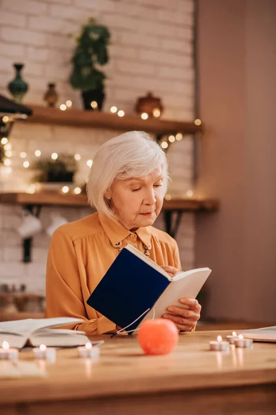 Ősz hajú, gyönyörű idős hölgy narancssárga blúzban könyvet olvas. — Stock Fotó
