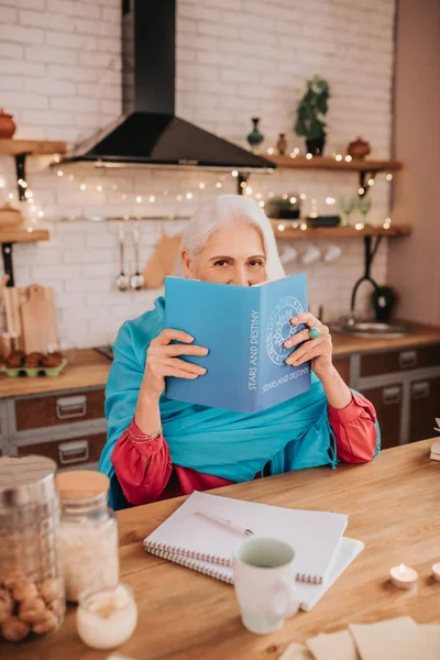 Ősz hajú, gyönyörű idős hölgy kék kendőben, becsukja az arcát egy könyvvel. — Stock Fotó