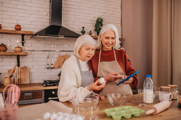 Damas sonrientes de pelo gris en delantales sintiéndose bien cocinando en la cocina — Foto de Stock