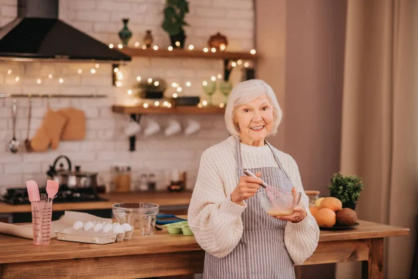 Cinzento de cabelos sorridentes senhora de pé perto da mesa na cozinha — Fotografia de Stock