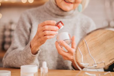 Grey-haired beautiful lady in grey blouse holding a jar of cream