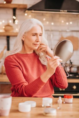 Grey-haired beautiful lady in red blouse looking at herself at the mirror