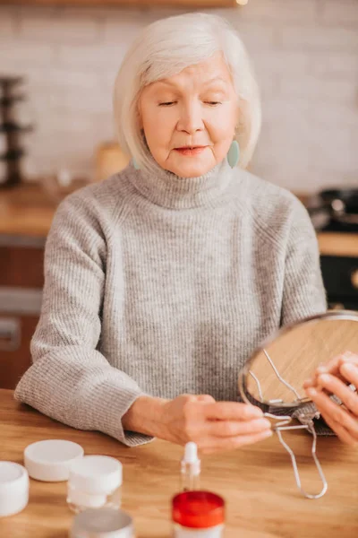 Grey-haired beautiful lady in grey blouse holding a mirror