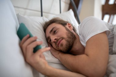 Uykucu adam yatağında telefonunu kullanıyor.