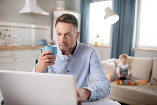 Homem barbudo em uma camisa azul olhando sério trabalhando em seu laptop — Fotografia de Stock
