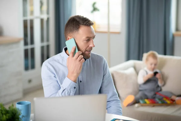 Vousatý muž v modré košili vypadající zapojeni, zatímco mluví po telefonu — Stock fotografie