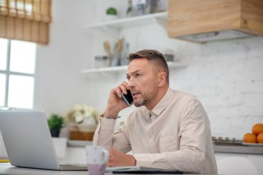 Yakışıklı sakallı adam mutfakta oturuyor ve bir telefon görüşmesi yapıyor.