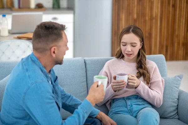 Дочка розмовляє з батьком, сидячи на дивані, дивлячись в чашку . — стокове фото