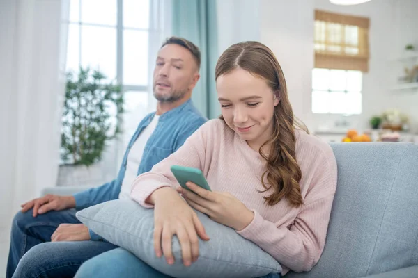 Daughter with smartphone looking away from father sitting on sofa. — Stock Photo, Image