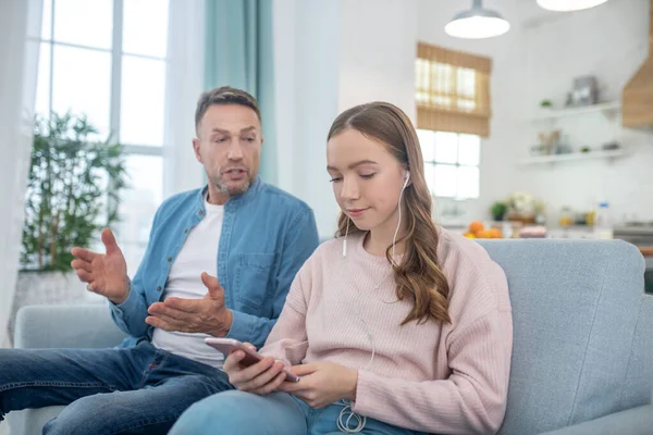 Daughter with smartphone, in headphones and father sitting on couch. — Stock Photo, Image