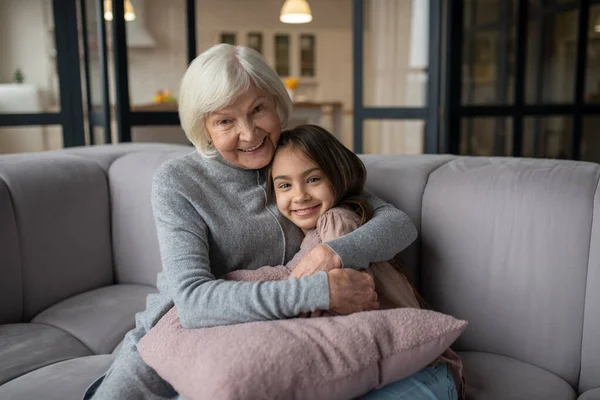 Großmutter und Enkelin umarmen sich und lächeln auf einem Sofa. — Stockfoto