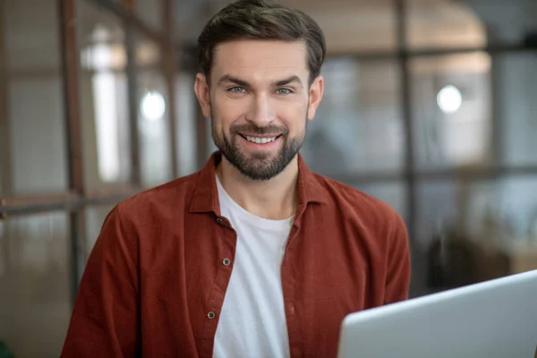 Jovem bonito em uma camisa marrom segurando um laptop e sorrindo — Fotografia de Stock
