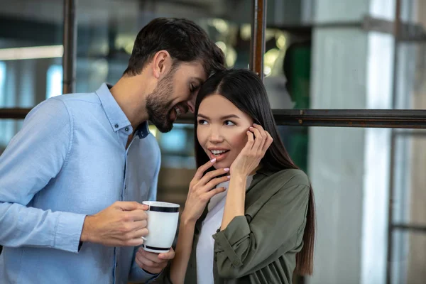 Tall young man having coffee and whispering something in the ear to his colleague